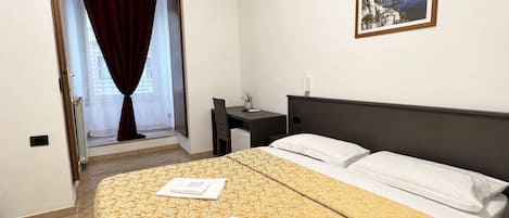 A hotel room with a bed, a desk, a chair, a window with curtains, and a framed picture on the wall.