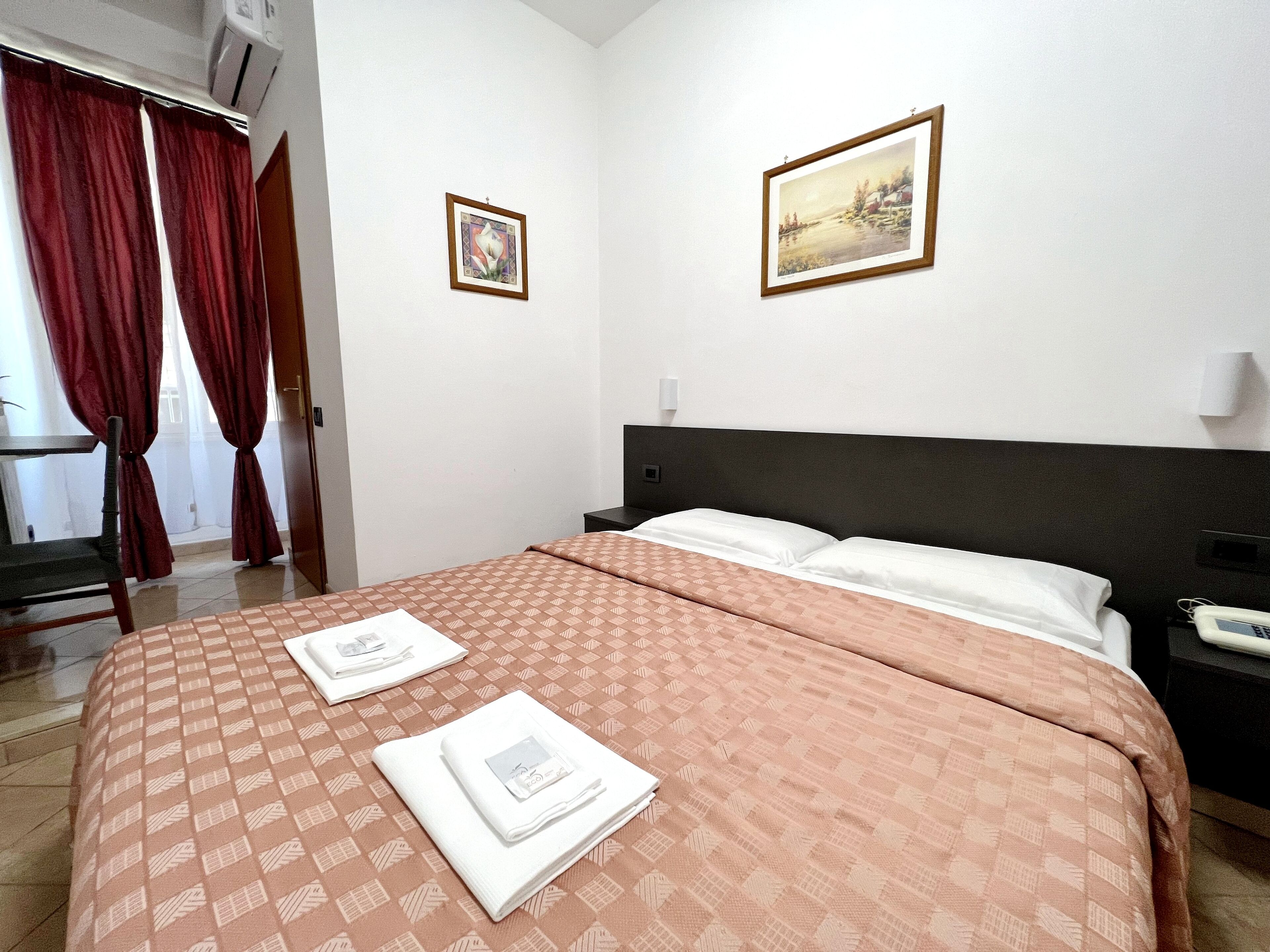 A hotel room with a large bed, two framed pictures on the wall, a bedside table with a phone, and a chair near the window with red curtains.
