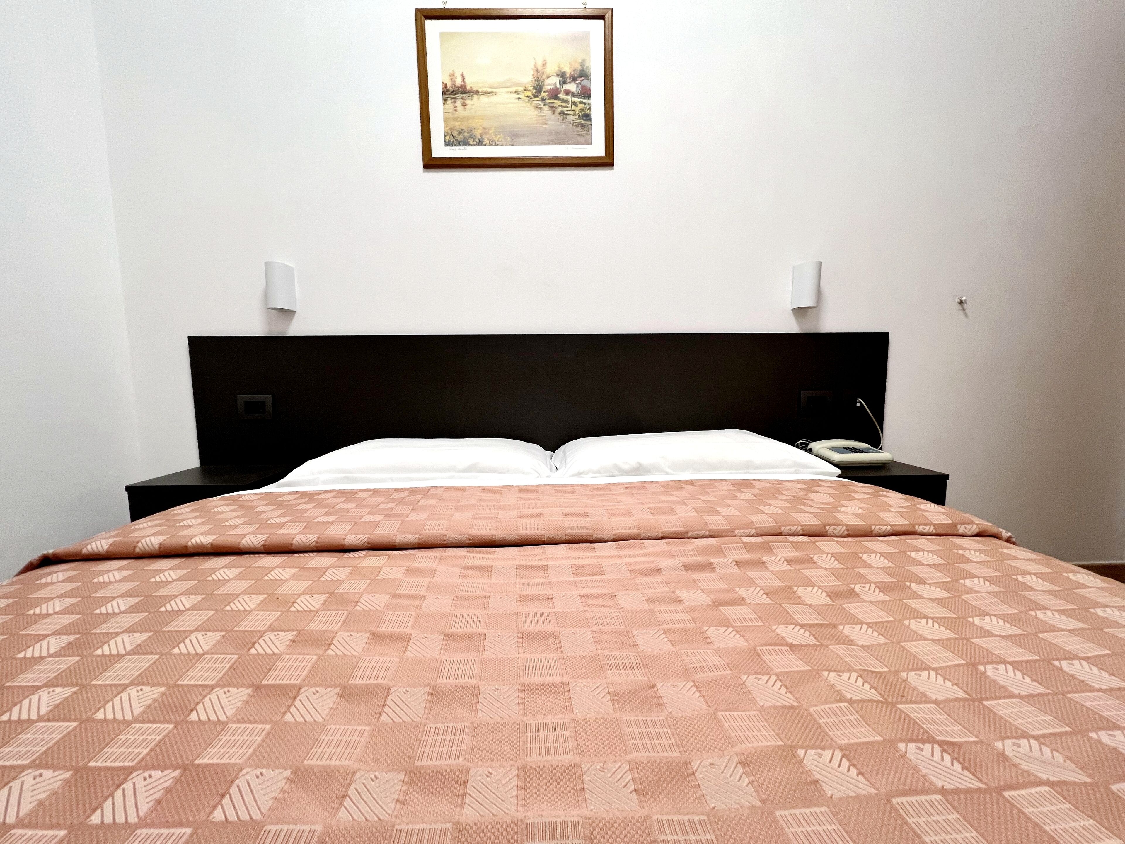 A hotel room with a bed, a nightstand, a bedside table, a lamp, and a framed picture on the wall.