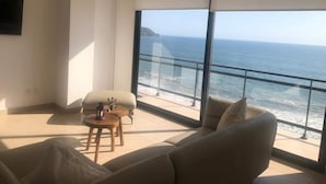 Un salón moderno con un amplio ventanal con vistas al océano, un sofá, una pequeña mesa con un jarrón y un televisor de pantalla plana montado en la pared.