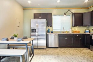 A kitchen with dark cabinets, a stainless steel refrigerator, a dishwasher, and a window with blinds.