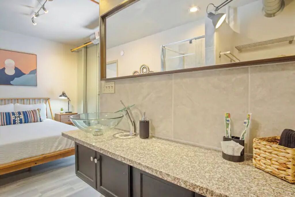 A modern bathroom with a large mirror, a glass bowl sink, and a bed with a wooden frame.