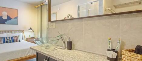 A modern bathroom with a large mirror, a glass bowl sink, and a bed with a wooden frame.