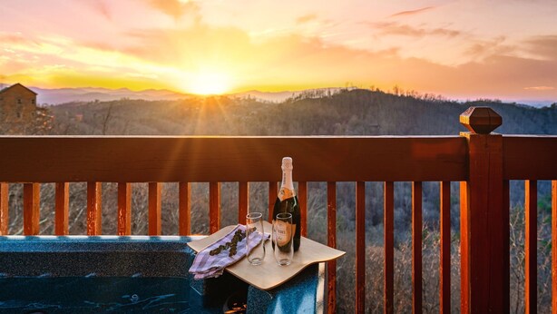 A wooden deck with a bottle of champagne and two glasses on a table, overlooking a mountainous landscape at sunset.