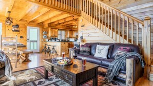 A cozy cabin interior with a wooden staircase, a dining area, a living room with a leather sofa, and a kitchen.
