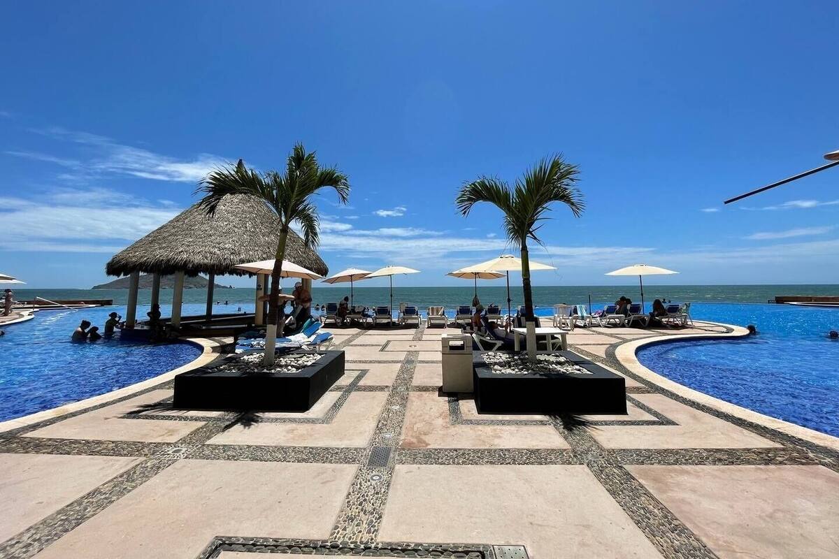 Área de piscina com espreguiçadeiras, guarda-sóis e uma estrutura com telhado de palha.