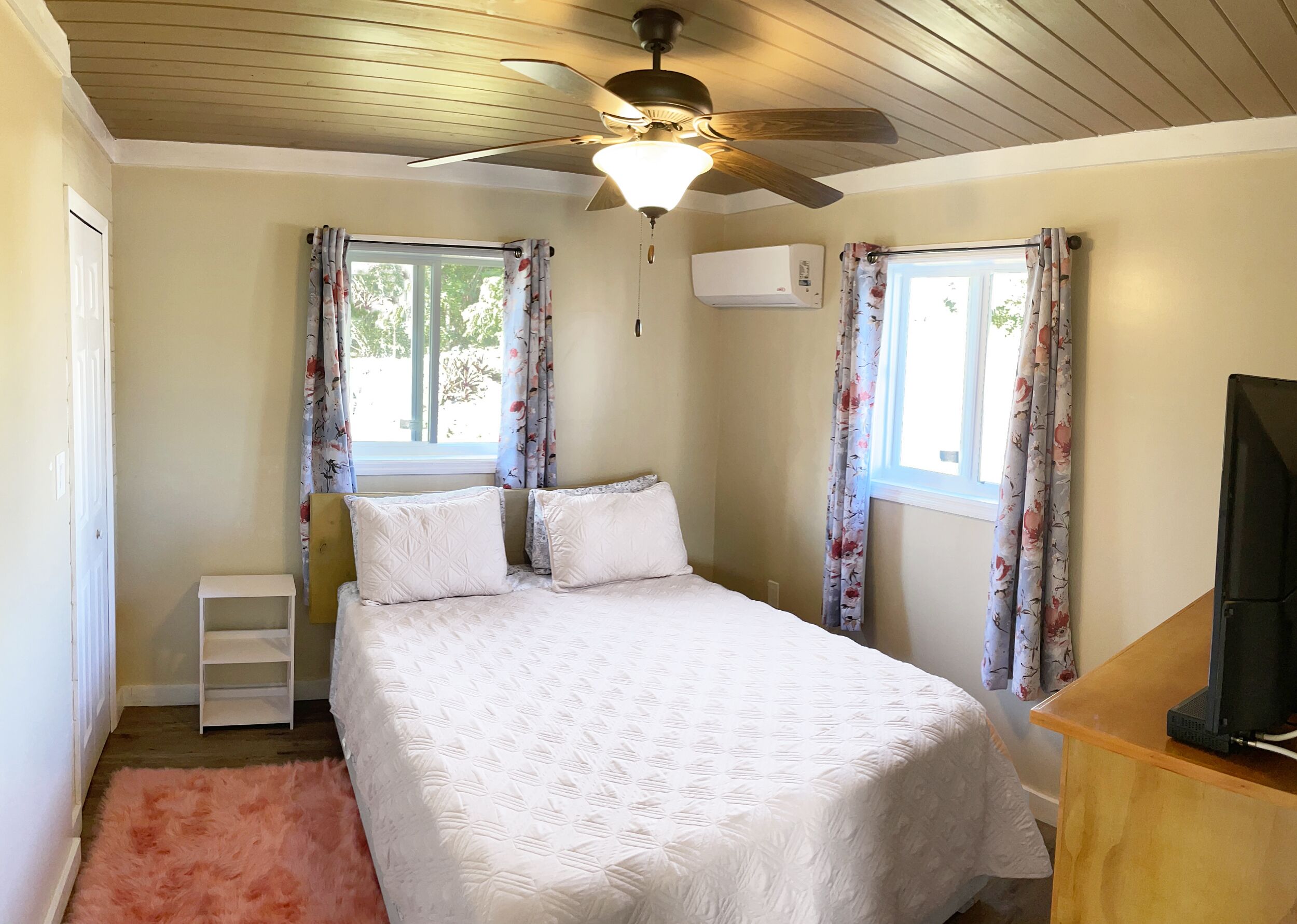 A bedroom with a bed, a ceiling fan, an air conditioning unit, and a television on a wooden table.