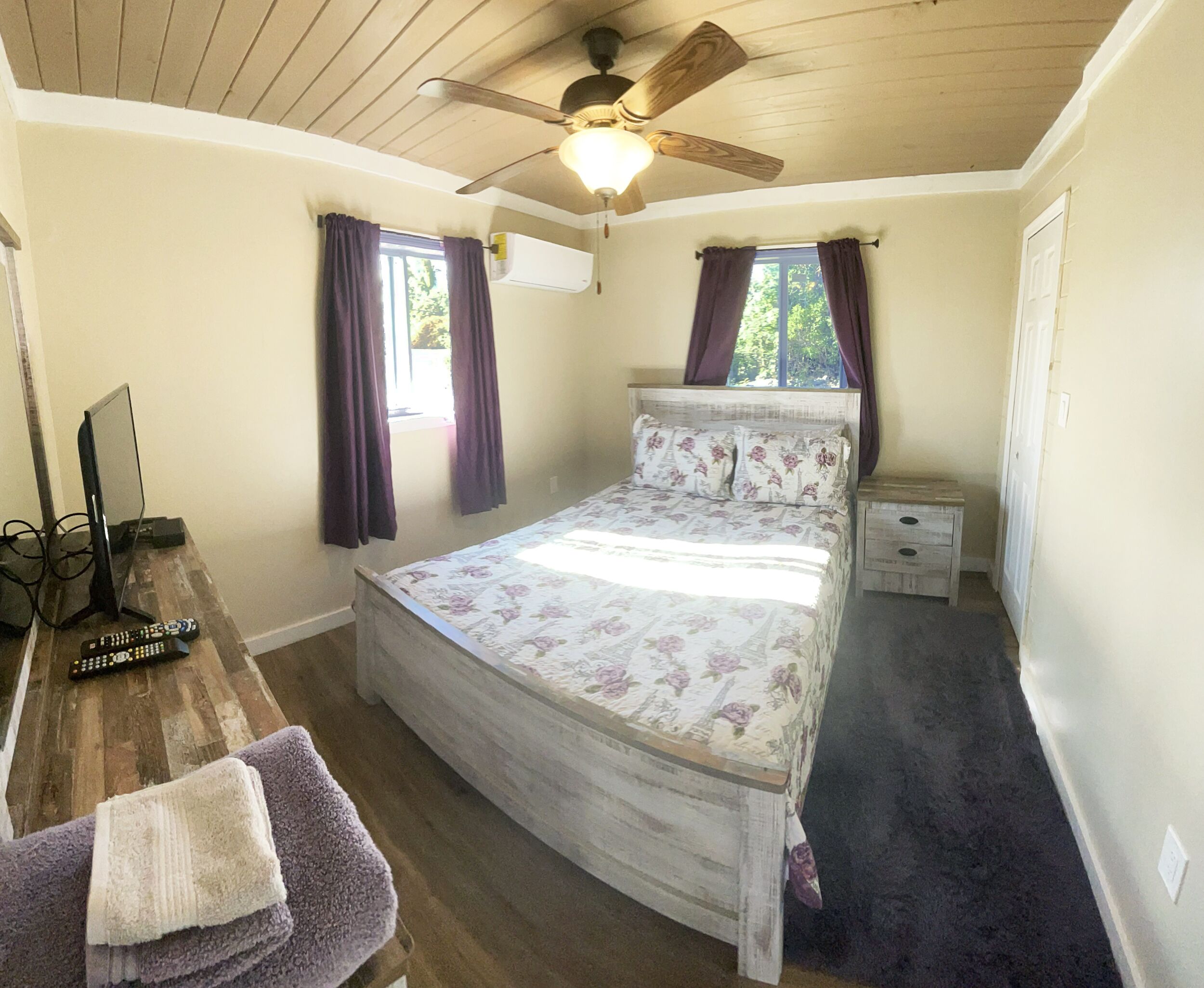 Ein Schlafzimmer mit Deckenventilator aus Holz, einem Bett mit floraler Bettwäsche, einem Nachttisch und einem Schreibtisch mit Fernseher und Fernbedienungen.