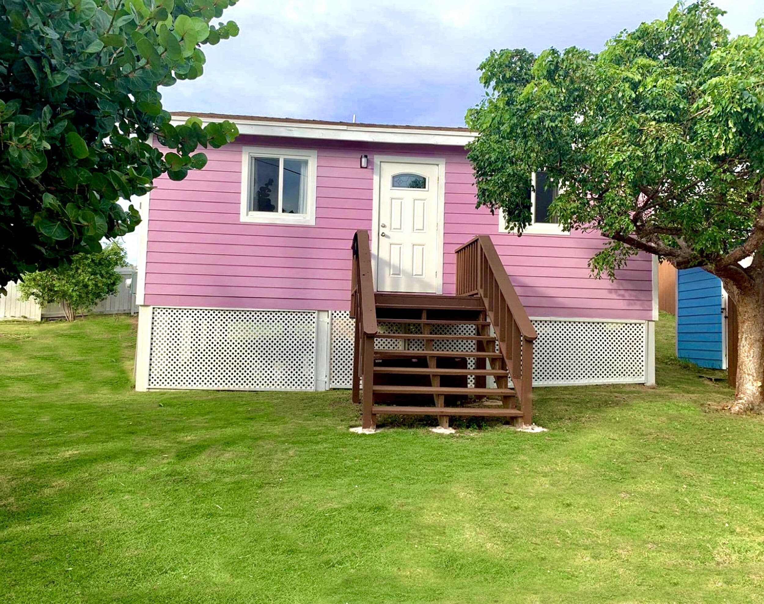 A pink house with a wooden staircase leading to a white door, surrounded by a white fence and a tree.