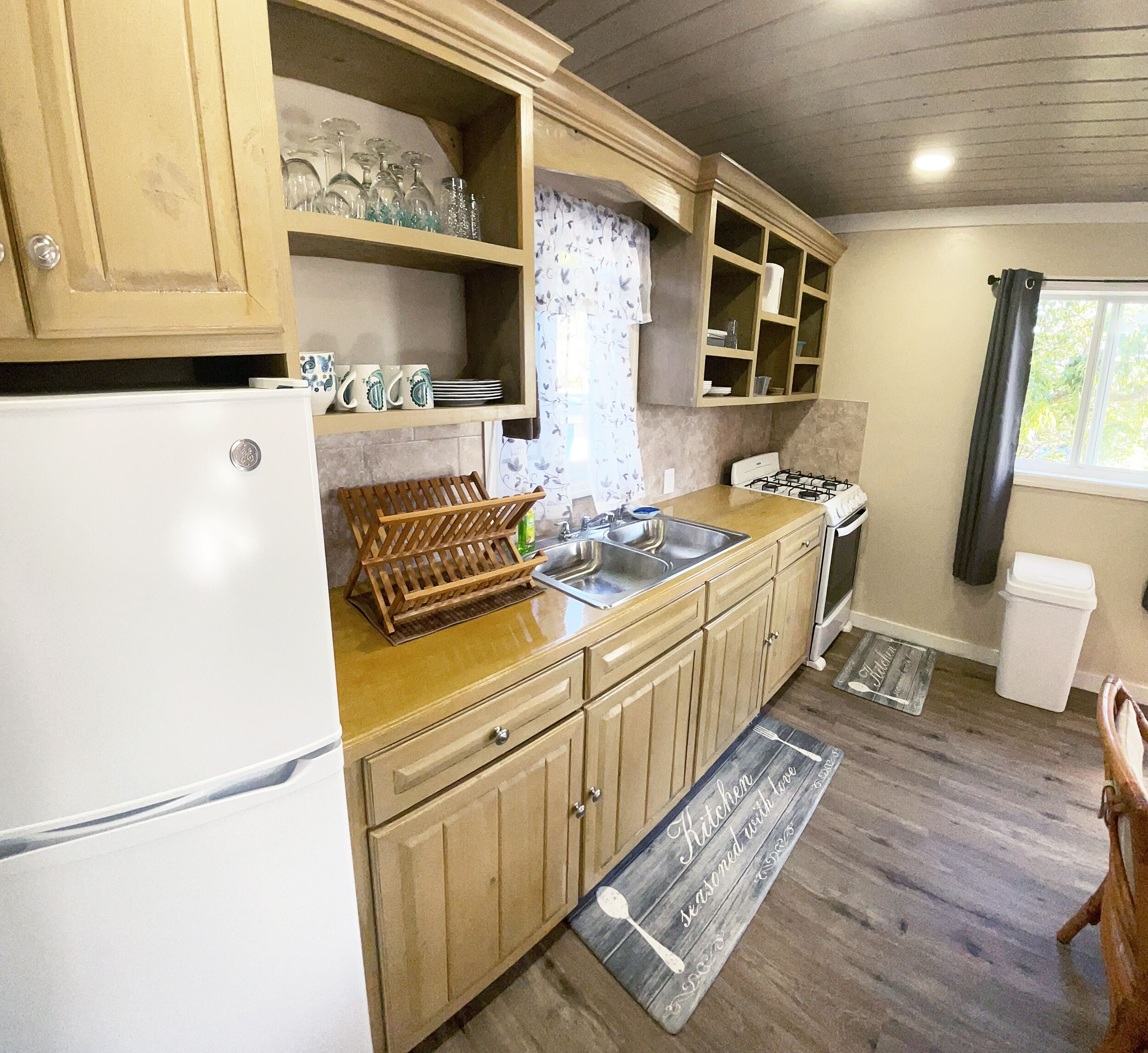 A compact kitchen with wooden cabinets, a white refrigerator, and a window with curtains.