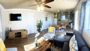 A spacious living area with a ceiling fan, a TV mounted on the wall, a dining table with chairs, and a kitchen in the background. - Cheerful Bahamian Pad. Walk to Beaches, Bars, Restaurants, Grocery-Liquor stores (hatchet bay)