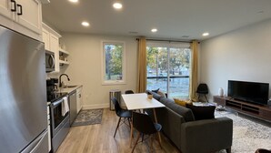 Cozinha moderna com frigorífico, fogão e micro-ondas. Possui uma área de jantar com mesa e cadeiras, e uma sala de estar com sofá e televisão.