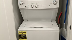 Máquina de lavar roupa com abertura frontal, painel de controle, etiqueta amarela de eficiência energética e um esfregão encostado na parede.