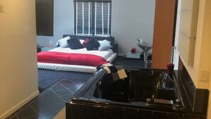 Une chambre d’hôtel avec une grande baignoire noire, un lit avec une literie rouge et blanche, et une fenêtre avec des stores.