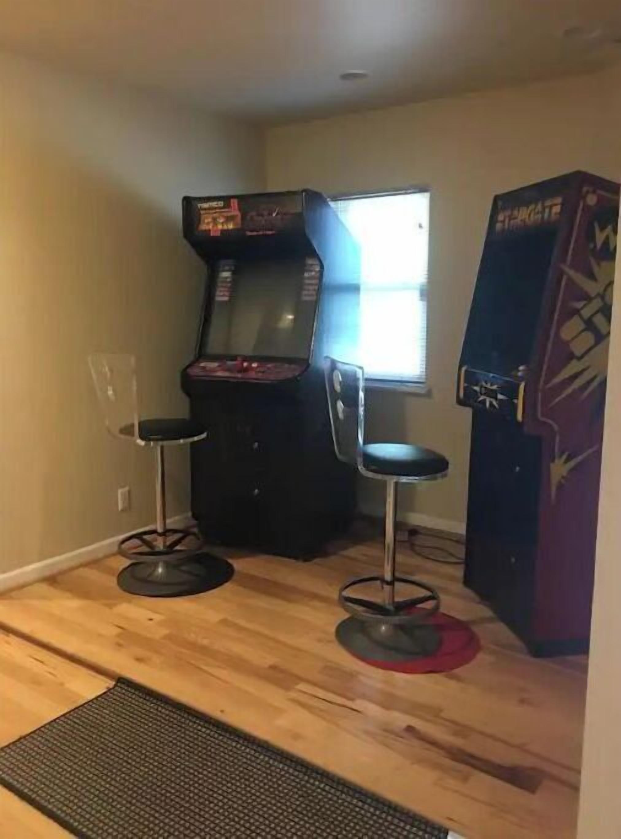 Ein Zimmer mit Holzboden, zwei Barhocker, einem großen Arcade-Automaten und einem Fenster.