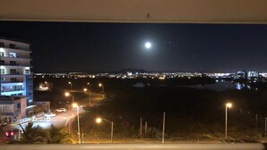 A nighttime view from a balcony overlooking a cityscape with a prominent moon in the sky, streetlights, and distant city lights reflecting on a body of water. - Departamentos MDR (Mazatlán)