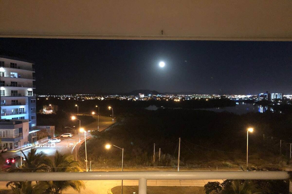 Vista nocturna desde un balcón con vistas a un paisaje urbano, destacando una luna grande en el cielo, farolas y luces de la ciudad que se reflejan en el agua.