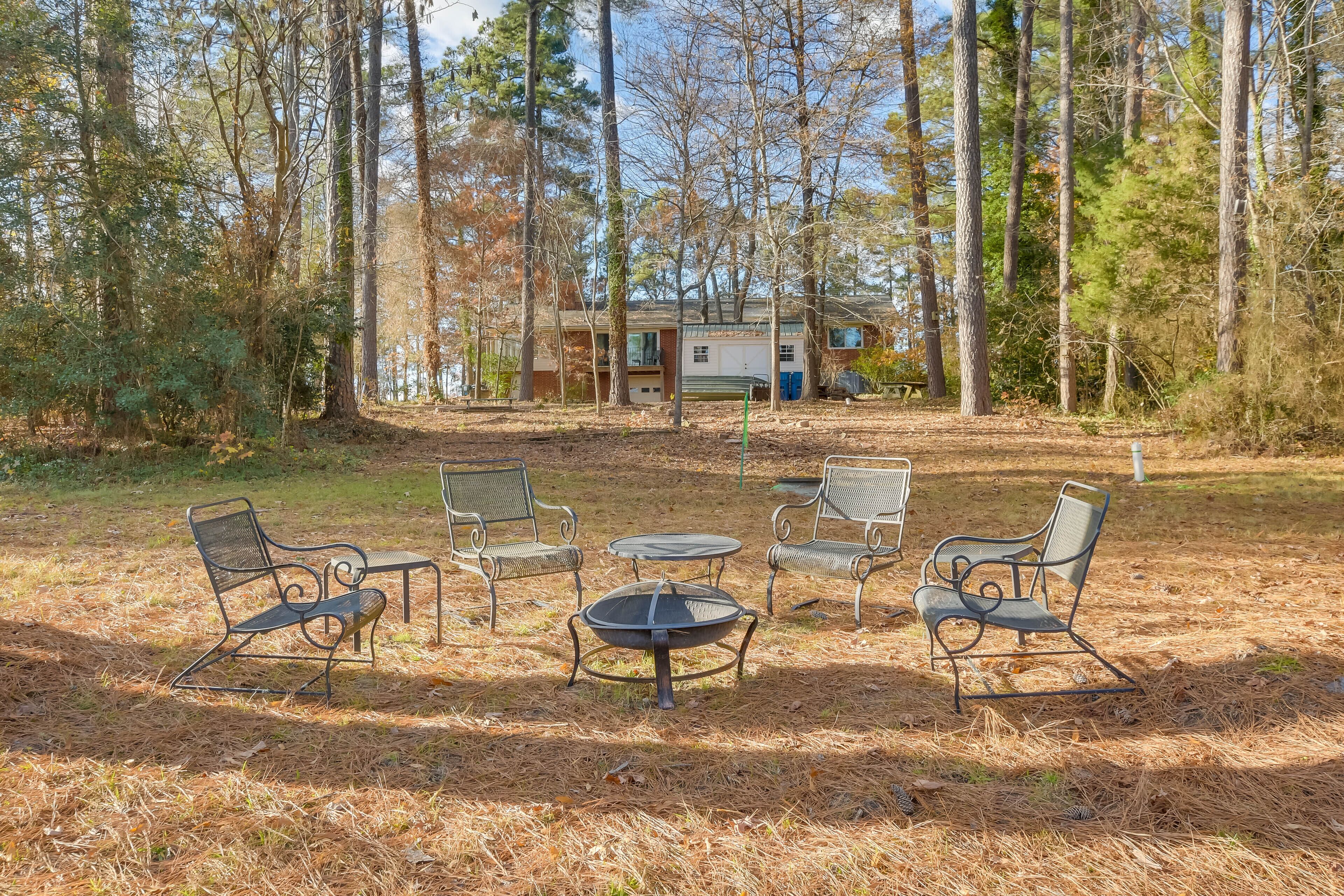 A campsite with metal chairs and a fire pit, surrounded by tall trees and a cabin in the background.