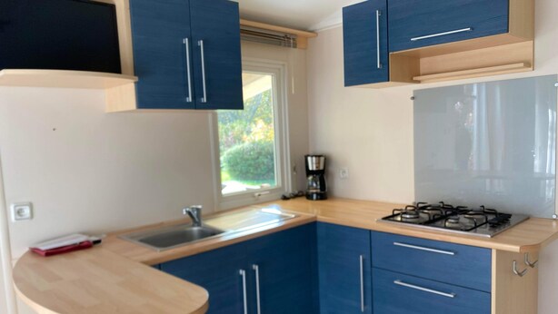 A compact kitchen with blue cabinets, a wooden countertop, a sink, a stove, and a window with a view of greenery. - Mobile home 6 persons. (Attigny)