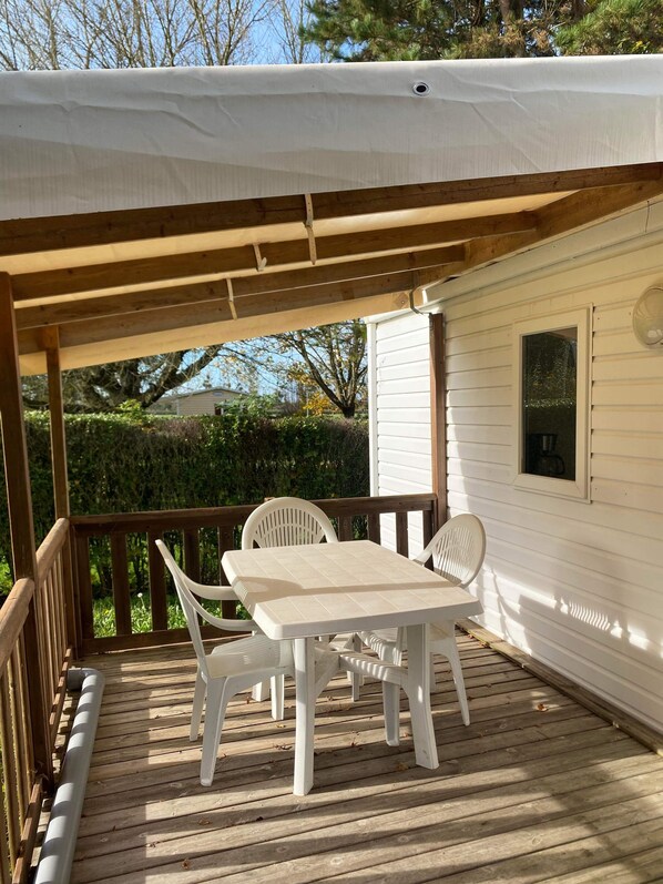 A covered outdoor deck with a table and chairs, a wooden railing, and a view of a white building with a window and a fan.