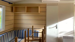 A room with a striped sofa, a wooden chair, a white air conditioning unit, and a window. - Mobile home 6 persons. (Attigny)