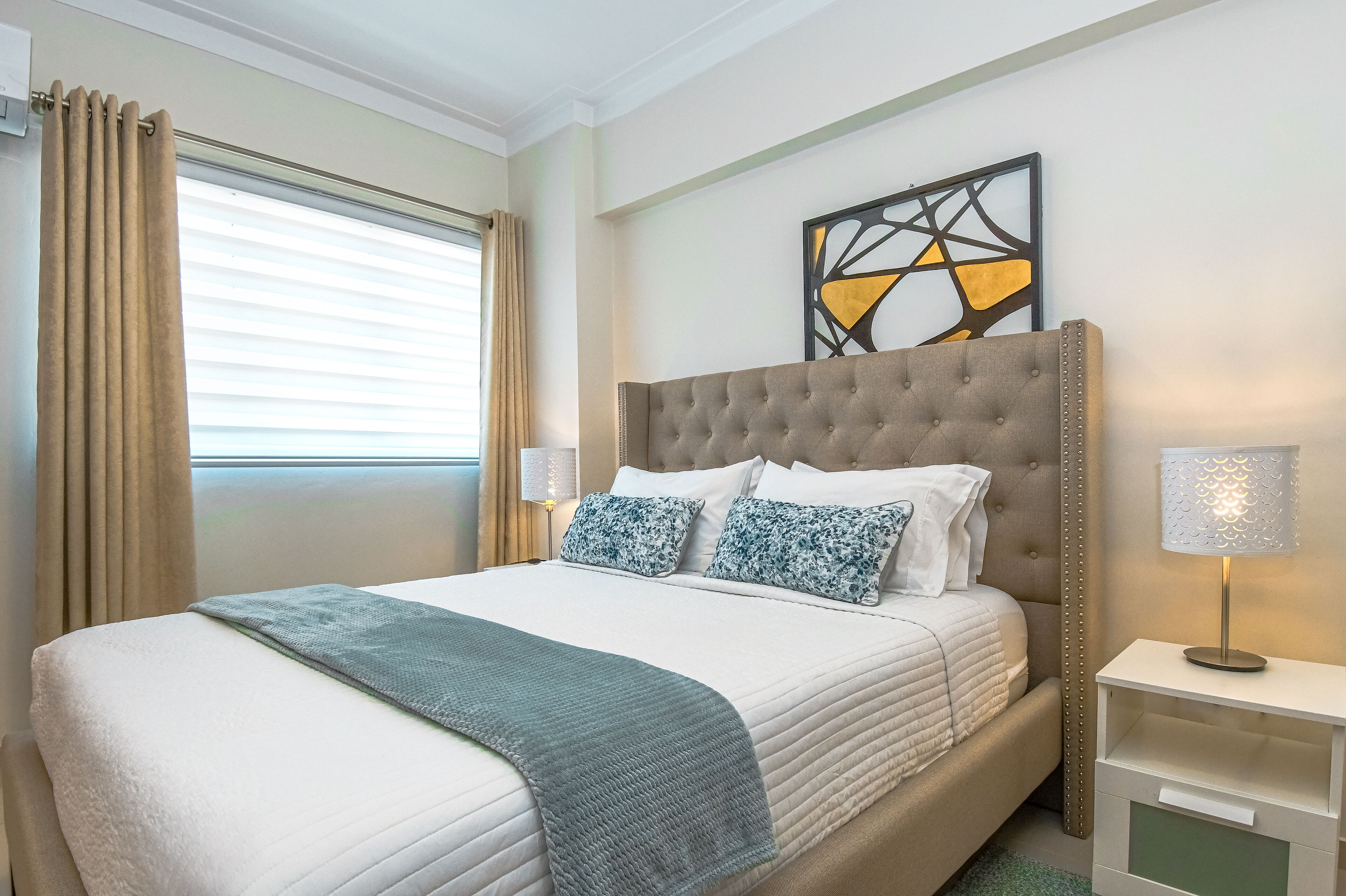 Una cama bien hecha con cabecera gris, ropa de cama blanca y una manta azul. Hay una mesita de noche con una lámpara y un adorno decorativo en la pared sobre la cama.