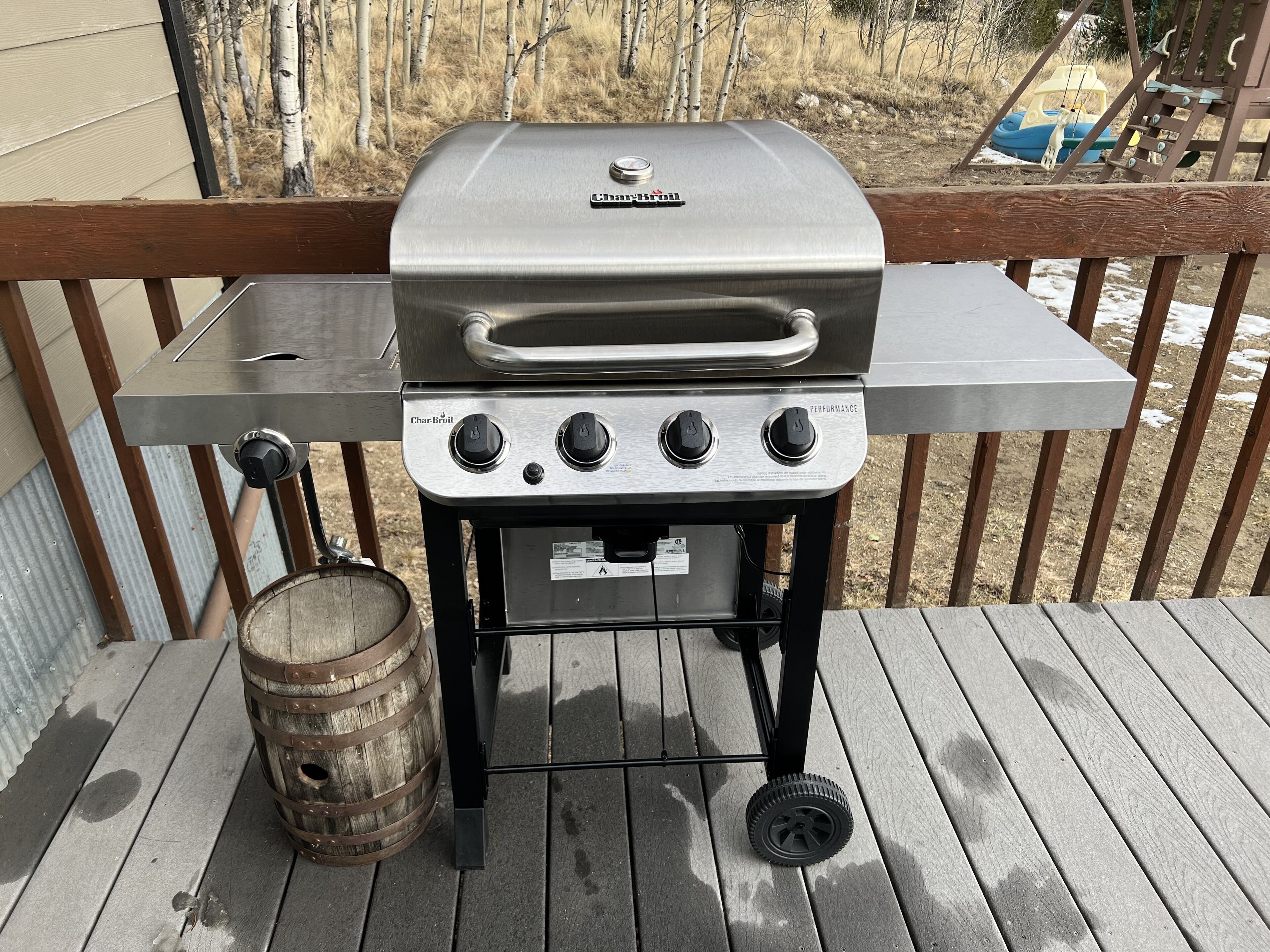 Ein Char-Broil Gasgrill mit vier Brennern und einer Seitenablage, platziert auf einer Holzterrasse, mit einem Fass in der Nähe.