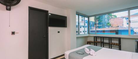 A bedroom with a bed, a TV mounted on the wall, a window with a view of buildings, and a ceiling fan.