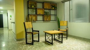 Un hall de hotel con piso de baldosas, dos sillas de madera, una mesita de madera y un estante para plantas en la pared.