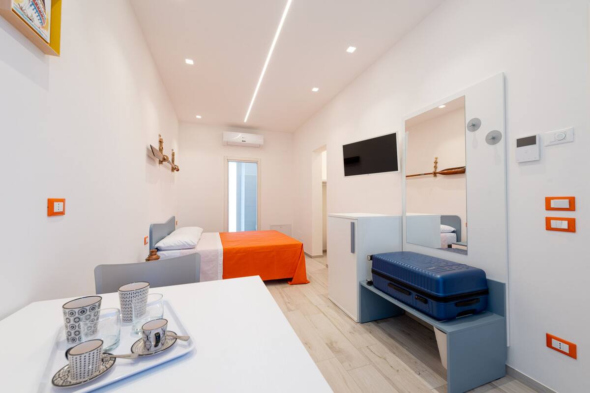 Une chambre d’hôtel moderne avec un lit, un bureau sur lequel se trouve un plateau avec des tasses, un téléviseur fixé au mur et une valise posée sur un banc.