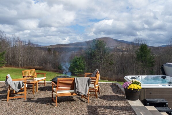 An outdoor seating area with wooden furniture and a hot tub, surrounded by a gravel patio and a scenic view of hills and trees.