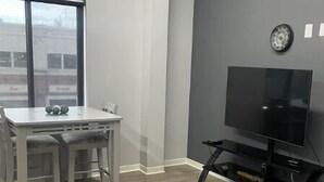 Een moderne woonkamer met een grijze muur, een flatscreen televisie op een zwart voetstuk, een eettafel met stoelen, en een groot raam met uitzicht op gebouwen.