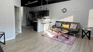 Een moderne woonkamer met een grijze bank, een salontafel en een vloerkleed. In de achtergrond is een keuken te zien met een magnetron en een koelkast.