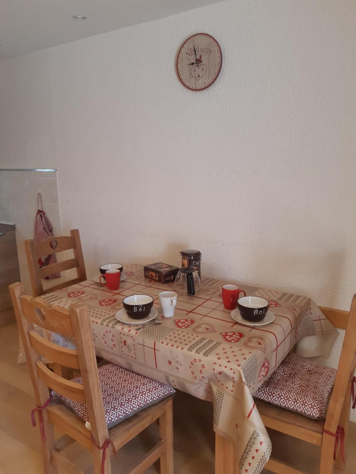 Un comedor con una mesa dispuesta para cuatro personas, con mantel estampado, sillas de madera y un reloj de pared.