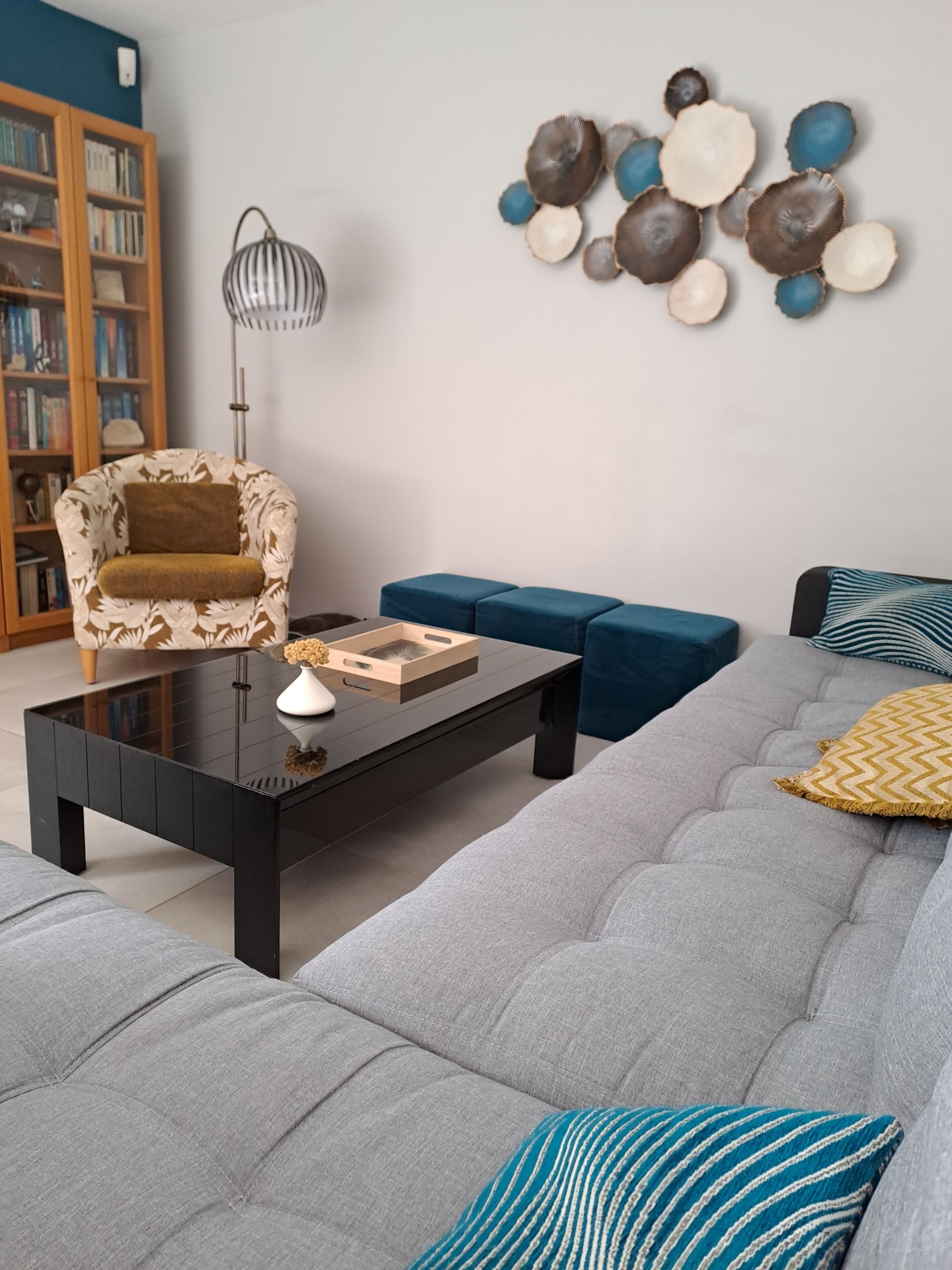 Een moderne woonkamer met een modulaire bank, een glazen salontafel, een fauteuil met een patroon en een decoratief object aan de muur.