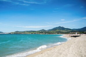 Une plage aux eaux turquoise et limpides, bordée d’un rivage sablonneux, avec des collines verdoyantes en arrière-plan.