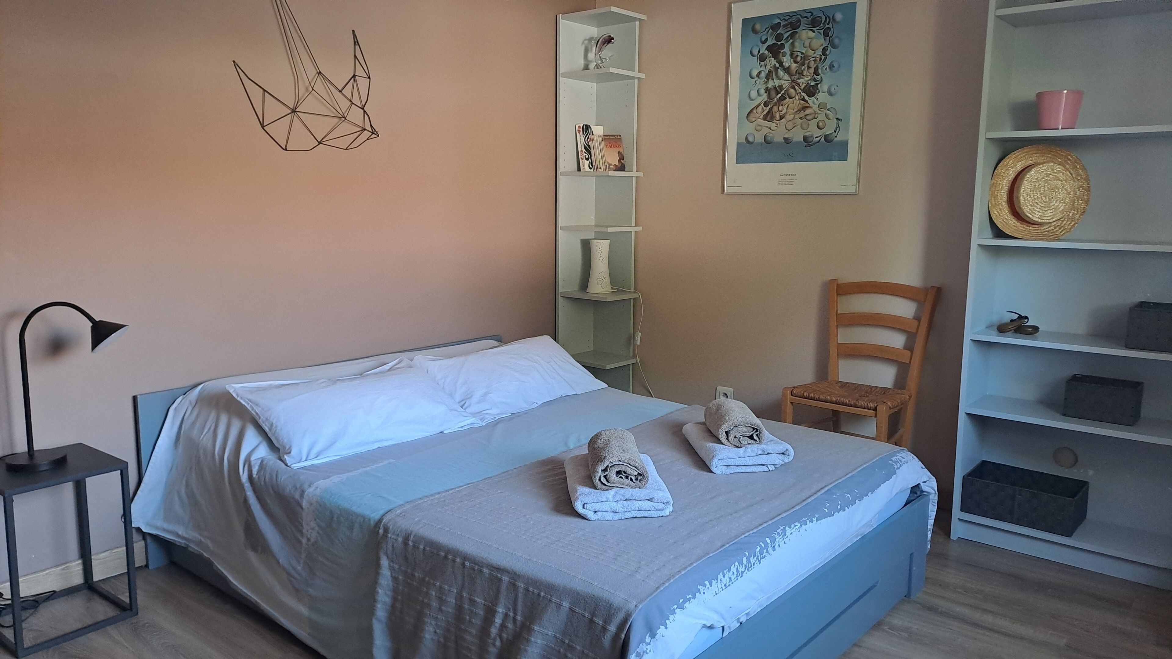 Une chambre à coucher bien rangée, avec un lit, une table de chevet, une chaise, une étagère ornée d’objets décoratifs et une œuvre d’art encadrée accrochée au mur.