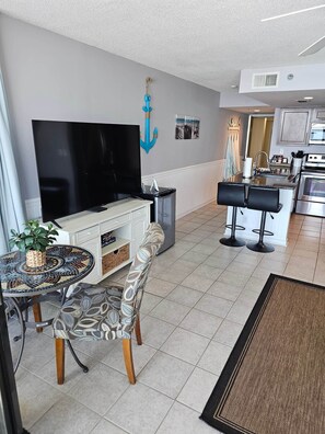 A modern living room with a flat-screen TV, a dining area with a patterned table and chairs, and a kitchen with a central island and bar stools. - Gulf-Front Condo, Balcony, 3 Pools, 1 Heated (Panama City Beach)