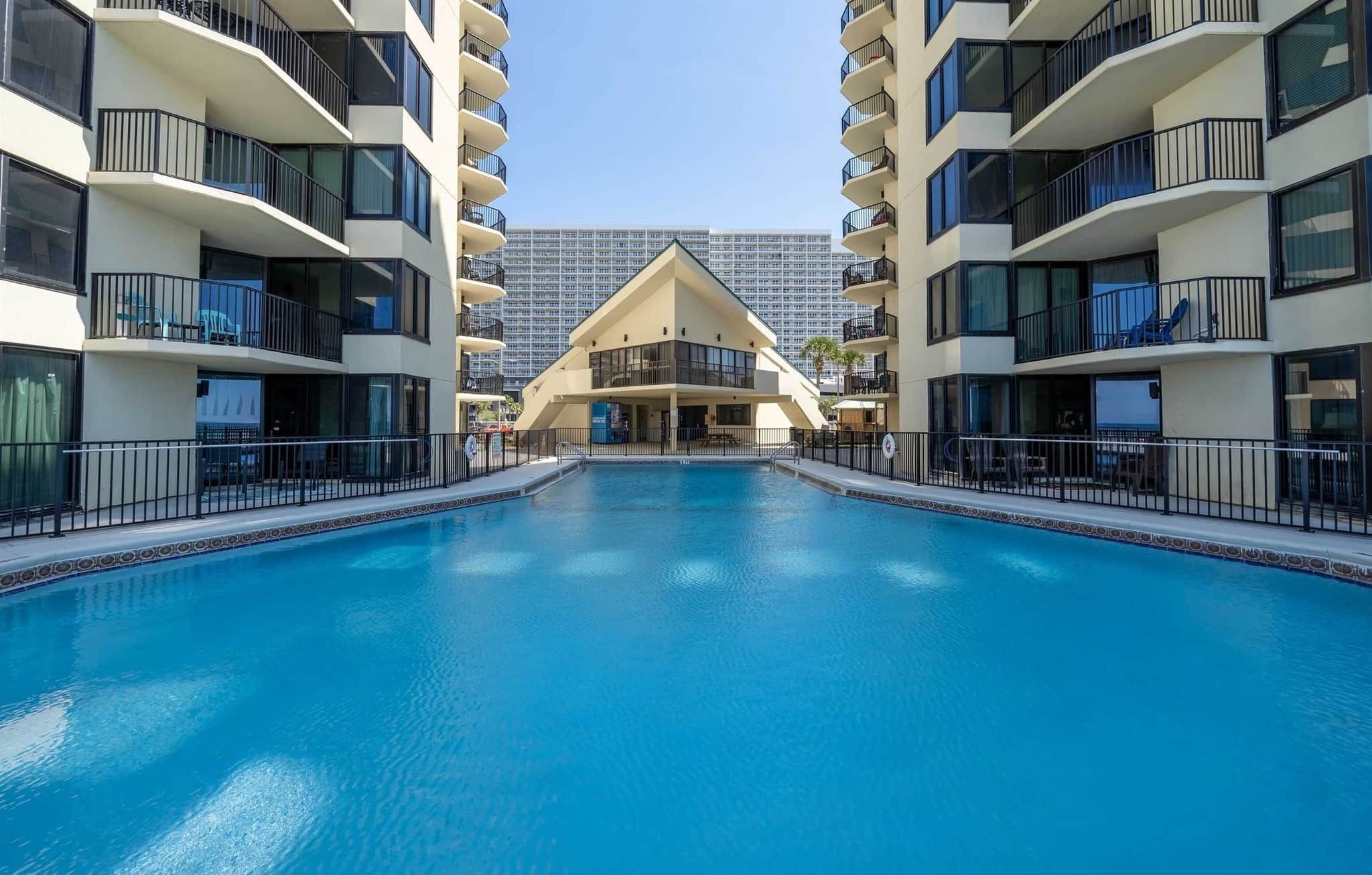 Una piscina rodeada de edificios modernos con balcones, un cielo despejado y un rascacielos a lo lejos.
