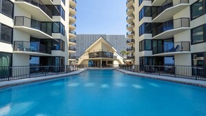 A swimming pool surrounded by modern buildings with balconies, a clear sky, and a distant high-rise building. - Gulf-Front Condo | Private Balcony, 3 Pools (1 Heated) (Panama City Beach)