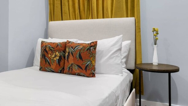 Ein Bett mit weißer Bettwäsche, ein einzelnes Kissen in Orange und Grün gemustert, sowie ein kleiner runder Tisch mit einer weißen Vase und gelben Blumen.