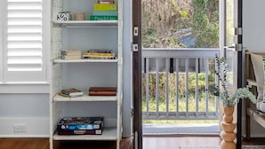 Ein Zimmer mit einem weißen Korbschrank, gefüllt mit Büchern und Spielen, einem Holzboden, einem Fenster mit Jalousien und Blick ins Grüne.