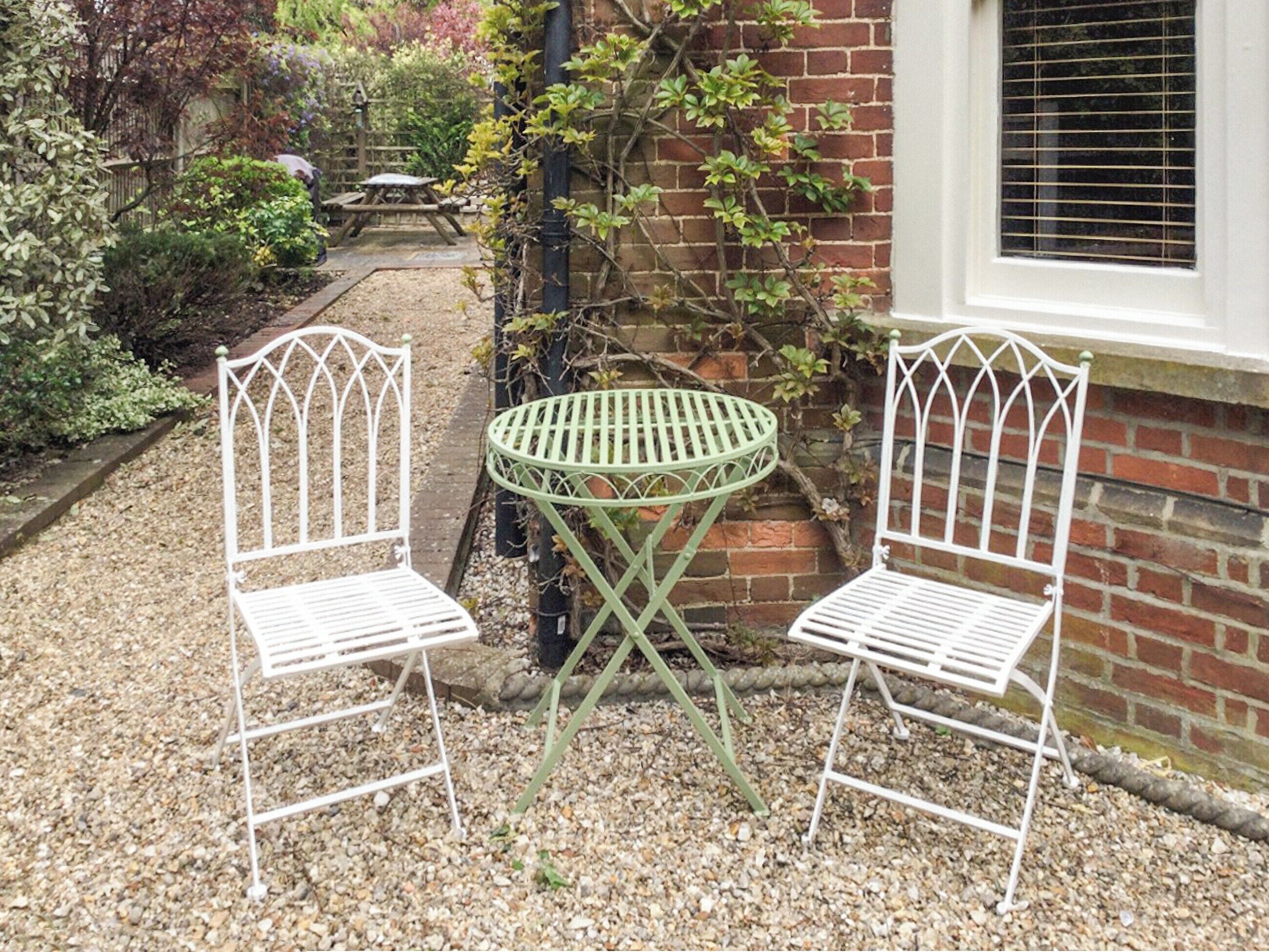 Two white metal chairs and a green metal table in a garden area with a brick wall and a window.