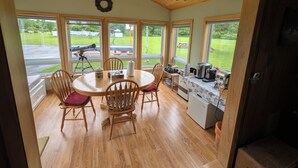 A cozy dining area with a round table, wooden chairs, and a view of a grassy yard through large windows. - Dog-Friendly large rural Estate just outside Alex Bay with hot tub and pool! (Alexandria Bay)