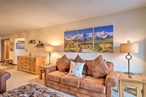 A cozy living room with a brown leather sofa, a wooden cabinet, and a coffee table. There are two wall-mounted lamps and a painting of mountains.