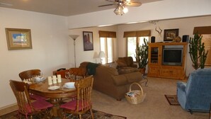 A dining area with a wooden table set for four, surrounded by chairs. There is a brown sofa and a blue armchair in the living room area. A television is mounted on a wooden cabinet, and a cactus plant is visible.