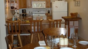 A kitchen with wooden cabinets, a stove, and a refrigerator. There are chairs and a dining table set with plates and glasses.