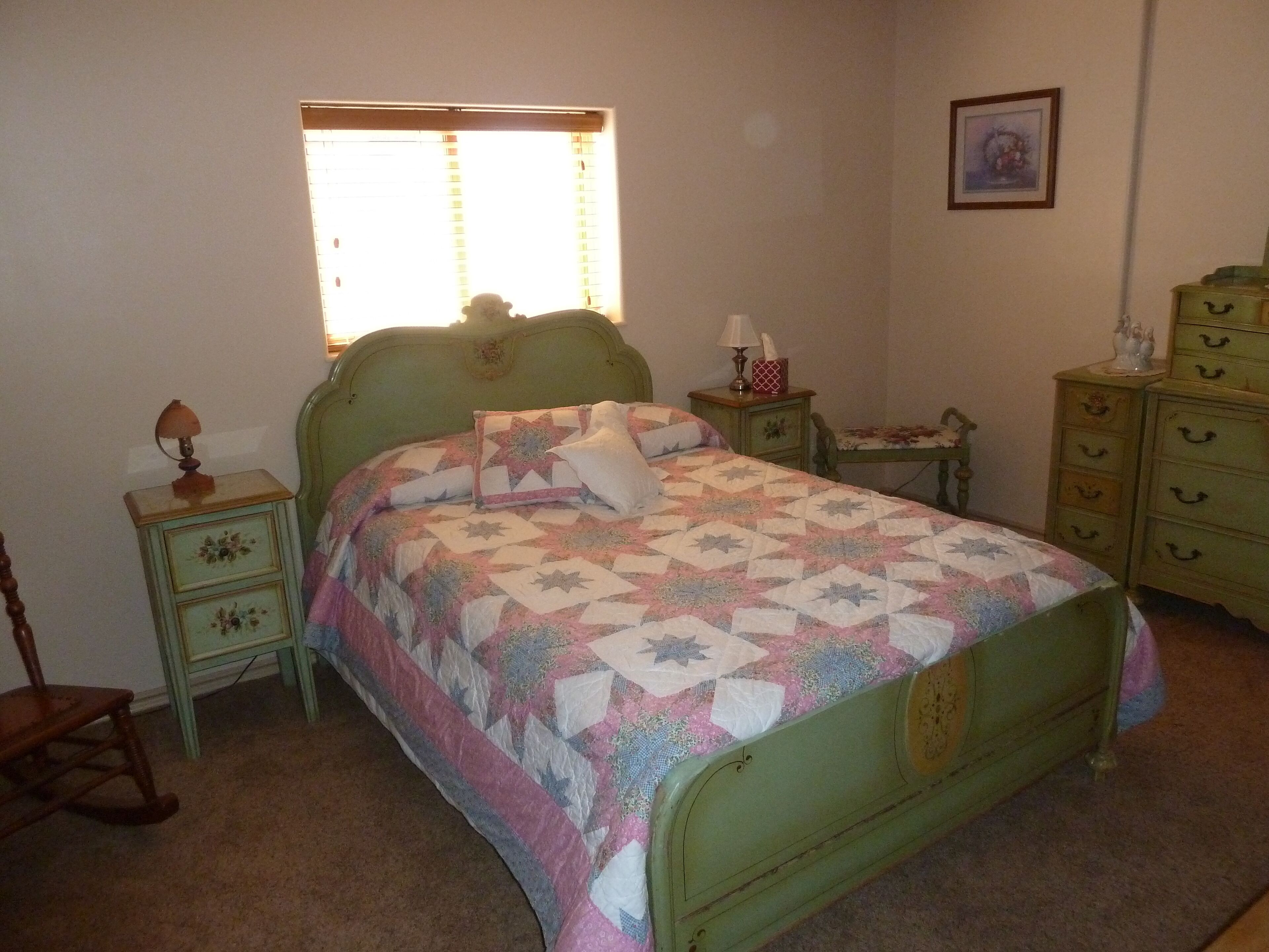 Un dormitorio con una cama verde, una mesita de noche con lámpara, un aparador y una ventana con persianas.