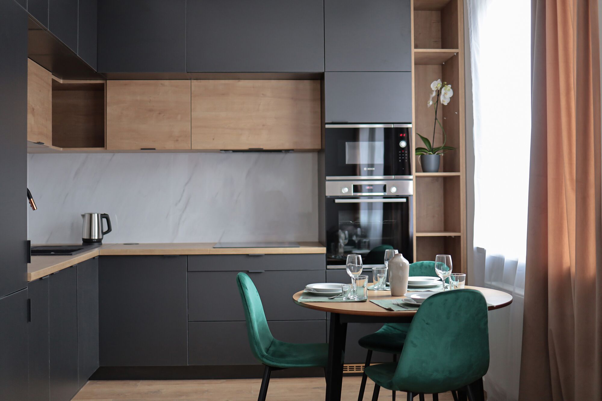 A modern kitchen with dark cabinetry, a wooden countertop, and a built-in oven. There is a dining table with chairs and a potted orchid on the windowsill.