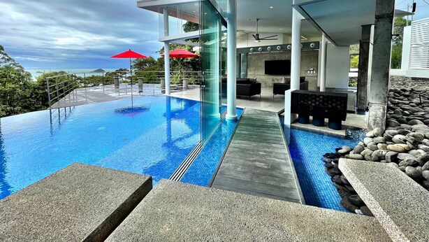 Una piscina infinita moderna con vistas despejadas al océano, un área de descanso cubierta con sombrillas rojas y una terraza con suelo de piedra.